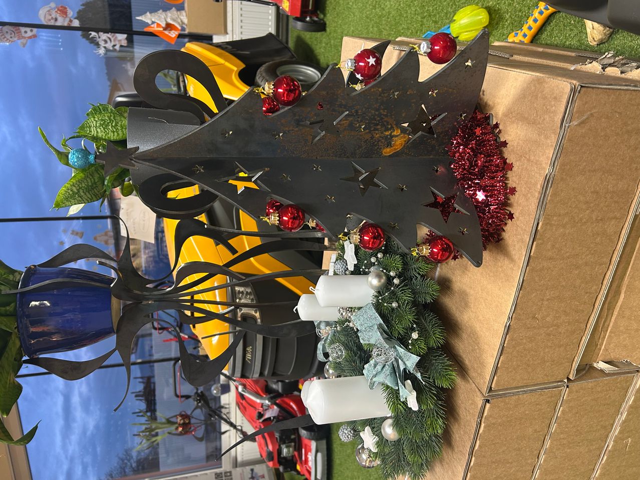 Ein Metall-Weihnachtsbaum mit roten Kugeln und Sternen, der auf einer braunen Pappschachtel ruht. Ein Kranz und Kerzen sind in der Nähe.