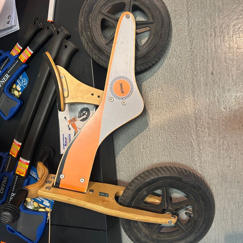 Ein zerlegter Kinderroller mit orangefarbenen und grauen Lenkern liegt auf einem Betonboden. Im Hintergrund befinden sich weitere Werkzeuge.