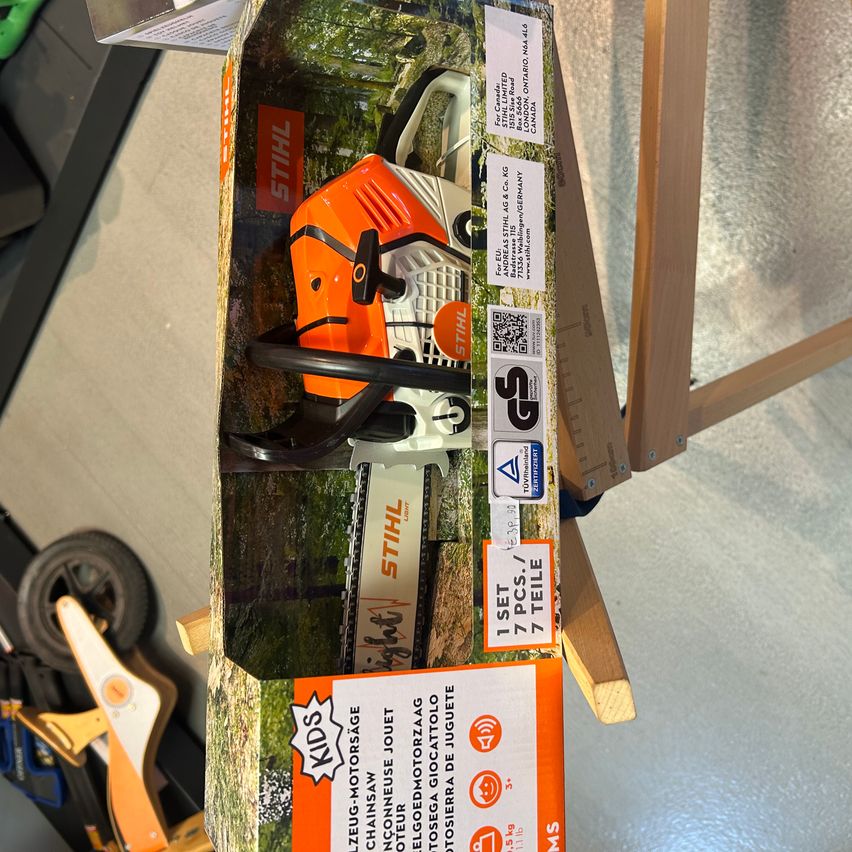 Eine Kettensäge mit orange und weißen Farben in einer geöffneten Schachtel mit Etikett. Sie hat verschiedene Zertifizierungen und einen Barcode. Die Schachtel trägt den Namen 'STIHL'.