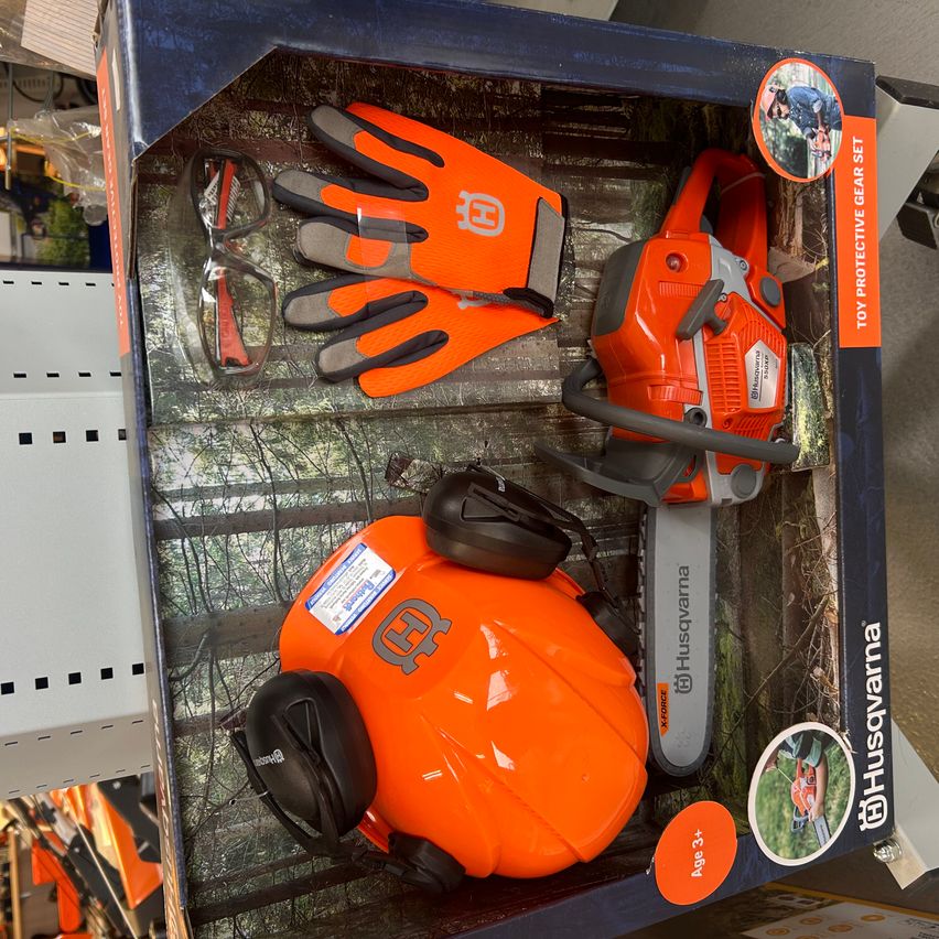 Eine blaue Schachtel enthält eine Spielzeugkettensäge, einen orangefarbenen Helm und orangefarbene Handschuhe. Die Kettensäge hat das Husqvarna-Logo und ist orange. Die Schachtel zeigt eine Waldszene.