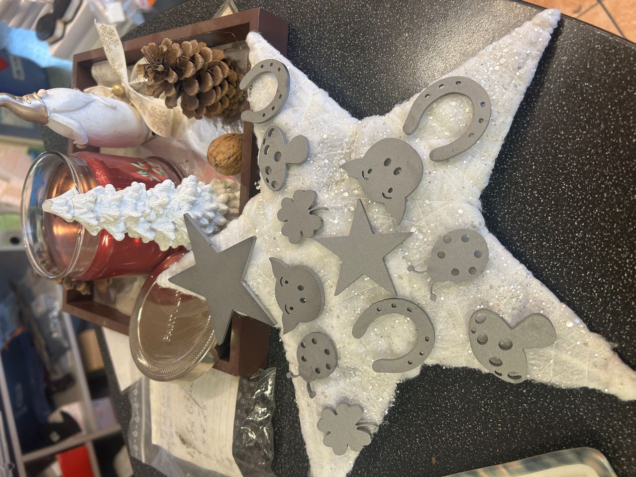 Ein sternförmiger Schmuck ist mit grauen Metallformen verziert, darunter Sterne, Hufeisen, Kleeblätter und ein Gesicht, auf einer schwarzen Oberfläche platziert.