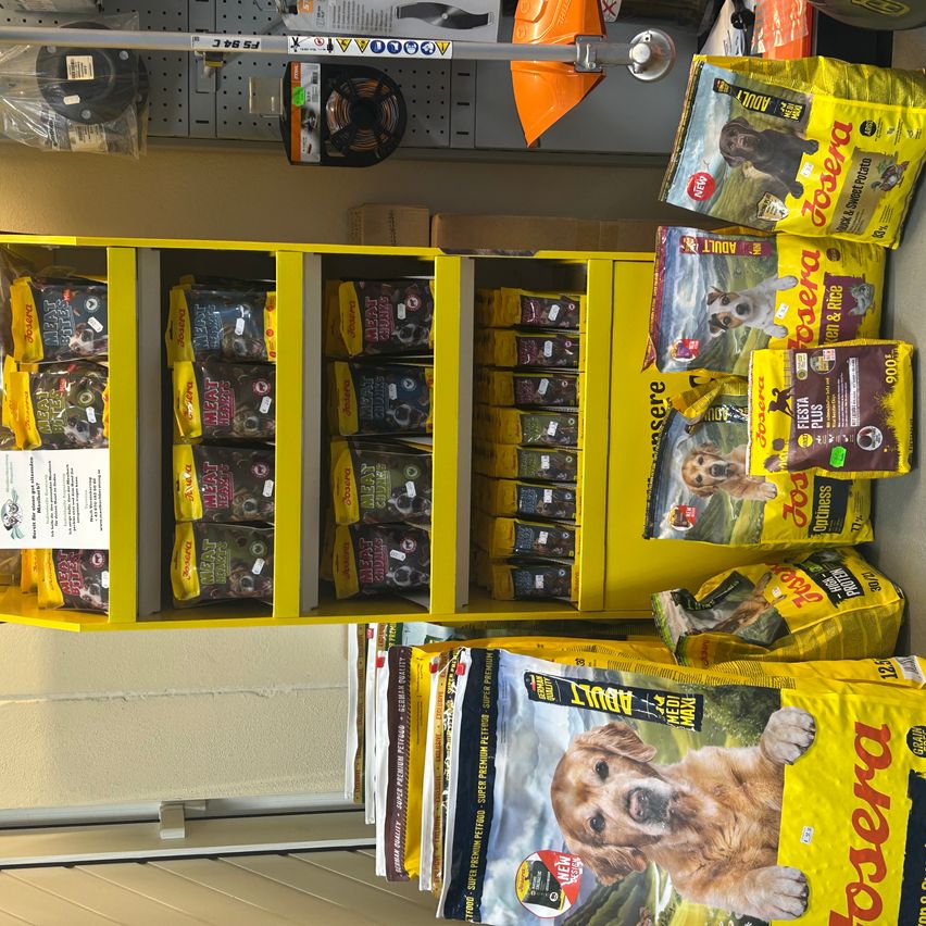 Ein gelbes Regal mit verschiedenen Hundefutterpackungen, darunter Marken wie Acana und Purina, in einem Geschäft ausgestellt. Rechts sind Säcke mit Hundefutter gestapelt.