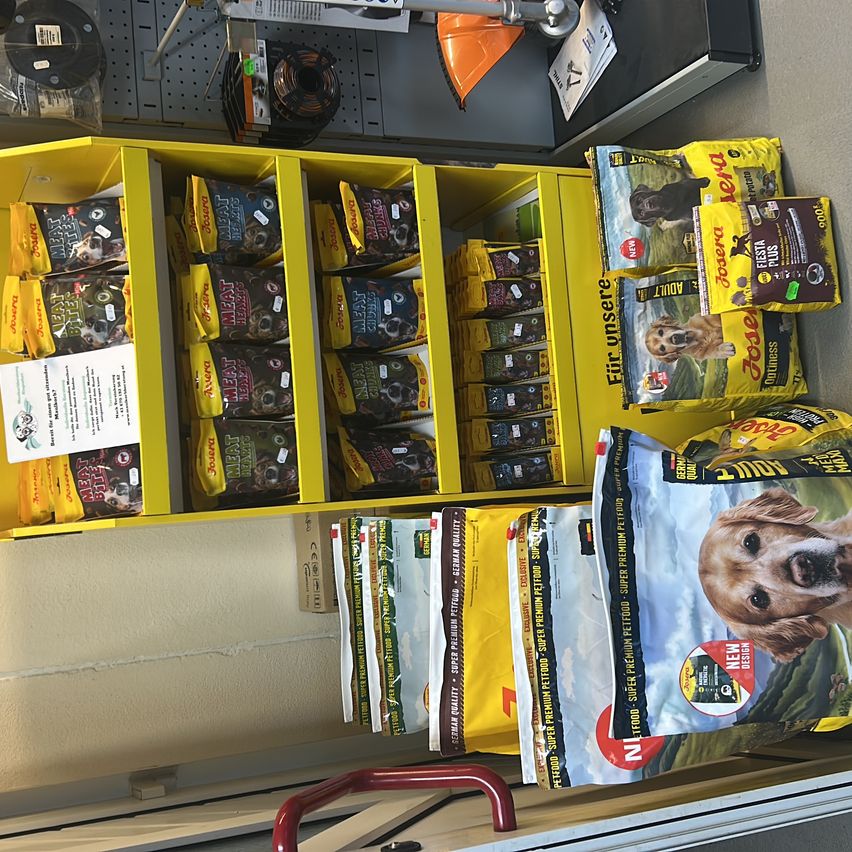 Ein gelbes Regal mit verschiedenen Packungen Hundefutter. Einige Packungen sind mit 'Hunde' und 'Fleischbissen' beschriftet. Es gibt auch mehrere Tüten mit Tierfutter, auf denen Hunde abgebildet sind.