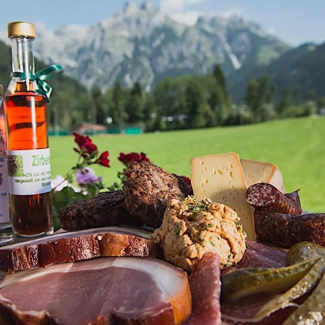 Ein Picknick-Arrangement mit verschiedenen Fleischsorten, Käse, Gurken und einer Flasche Sirup. Im Hintergrund befinden sich Berge und ein Feld.