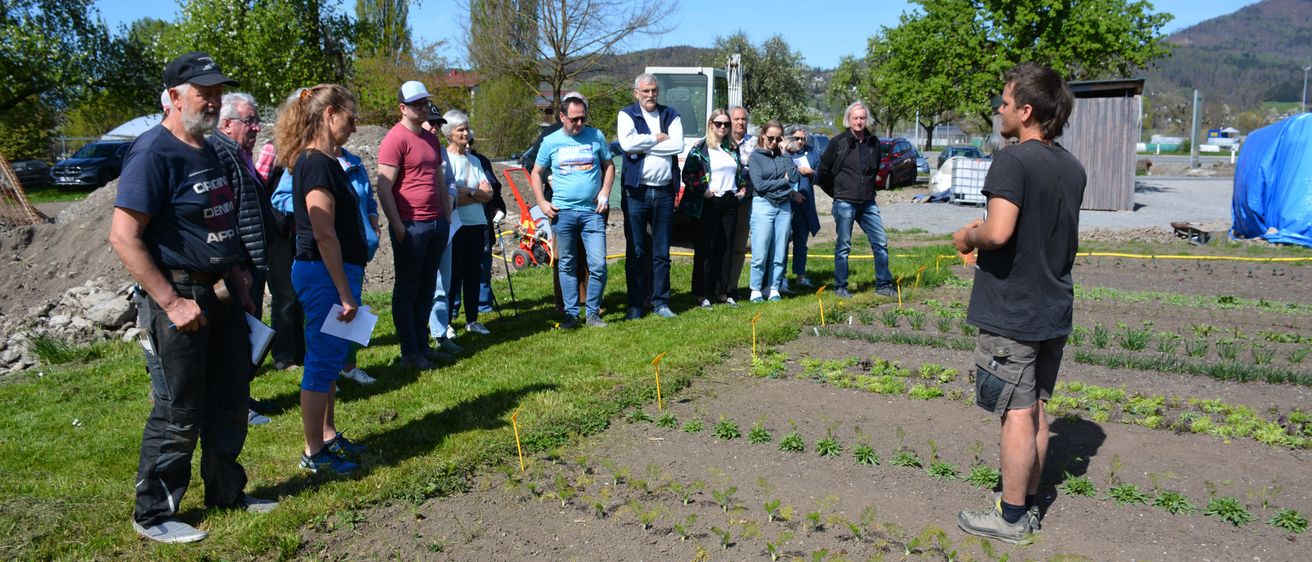 Eine Gruppe von Menschen steht in einem Garten mit grünem und braunem Boden und hört einem Mann vor ihnen zu.