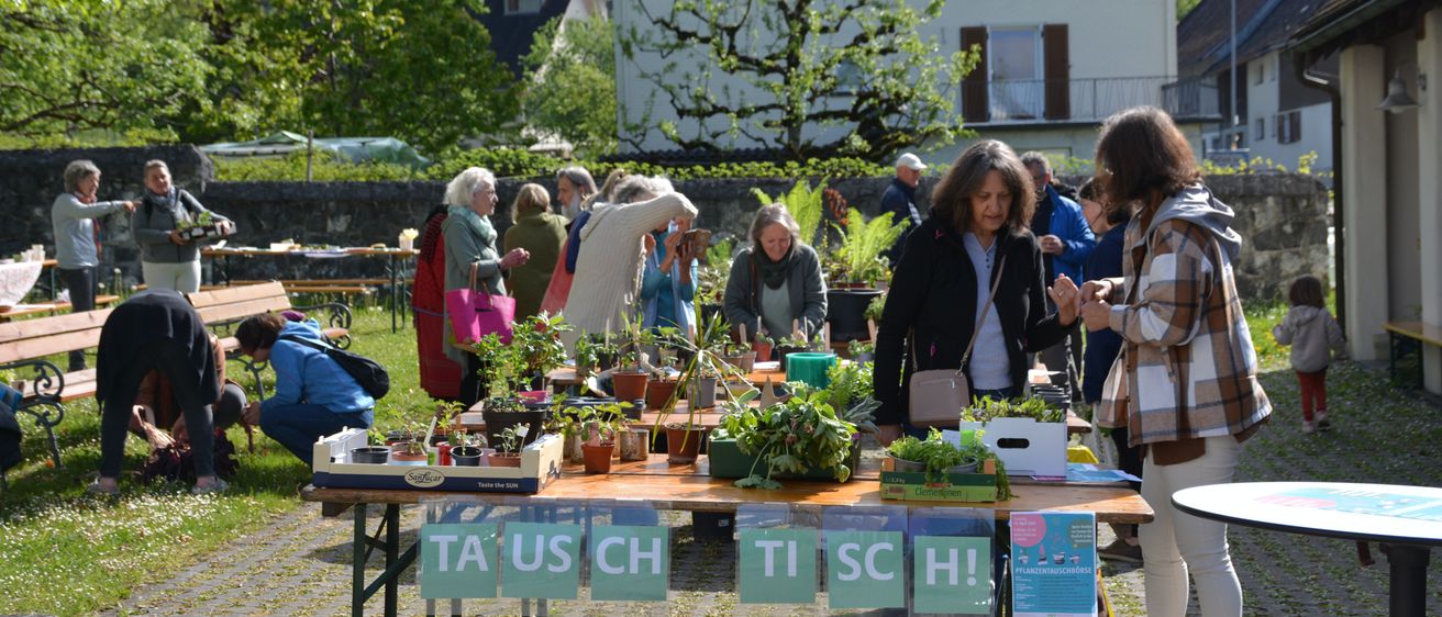 Ein Tisch im Freien mit Topfpflanzen und um ihn herum versammelten Menschen. Eine Frau steht vor dem Tisch. Der Tisch hat grüne Buchstaben, die das Wort Tausch bilden. Hinter dem Tisch steht ein weißes Gebäude.