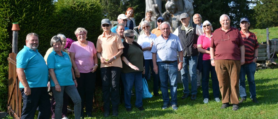 Eine Gruppe von Männern und Frauen steht im Halbkreis auf einem Rasenfeld mit einer großen Skulptur dahinter. Sie tragen Freizeitkleidung und einige von ihnen tragen eine Brille.