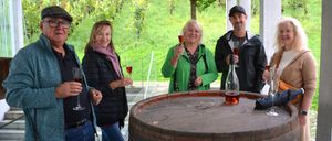 Fünf Personen stehen neben einem Fass. Jeder hält ein Weinglas. Eine Flasche Wein steht auf dem Fass. Sie stehen auf einer Terrasse mit einem Weinberg im Hintergrund.