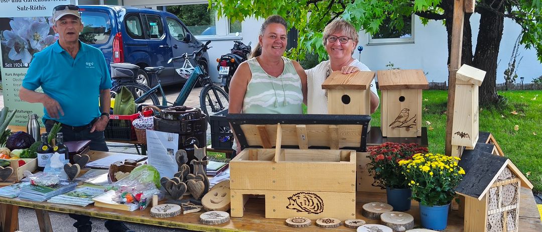 Drei Menschen stehen lächelnd unter einem Zelt neben einem Tisch mit Vogelhäusern, Blumen und anderen Gegenständen. Ein Mann trägt einen Hut und Turnschuhe, und eine Frau trägt eine Brille. Im Hintergrund stehen ein Van, Fahrräder und ein Baum.