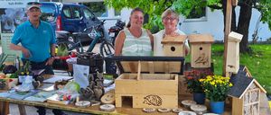 Drei Menschen stehen lächelnd unter einem Zelt neben einem Tisch mit Vogelhäusern, Blumen und anderen Gegenständen. Ein Mann trägt einen Hut und Turnschuhe, und eine Frau trägt eine Brille. Im Hintergrund stehen ein Van, Fahrräder und ein Baum.