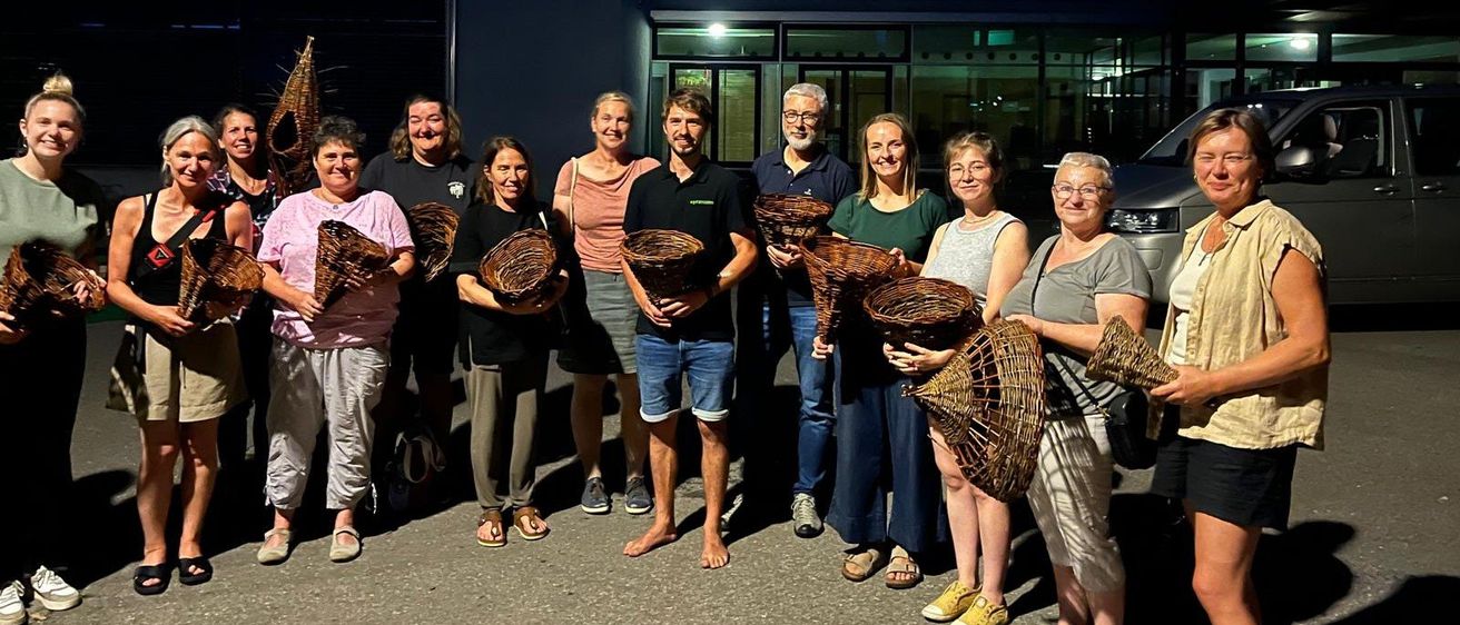 Eine Gruppe von Menschen, sowohl Männer als auch Frauen, stehen nachts draußen und halten Weidenkörbe in der Hand, lächeln für die Kamera. Ein Gebäude mit Glasfenstern ist hinter ihnen.