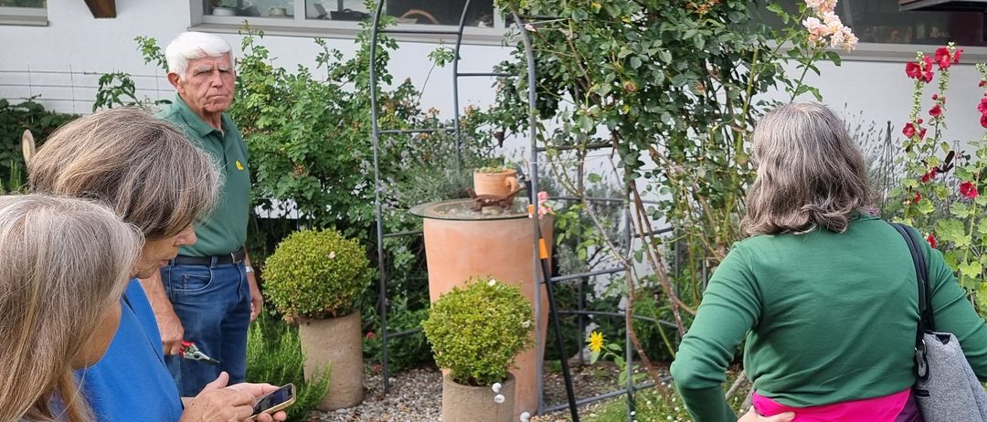 Eine Gruppe von Menschen ist draußen in einem Garten. Eine Person hält ein Telefon und schaut nach unten. Ein älterer Mann steht neben einer Frau, die die Hände auf die Hüften gestemmt hat.