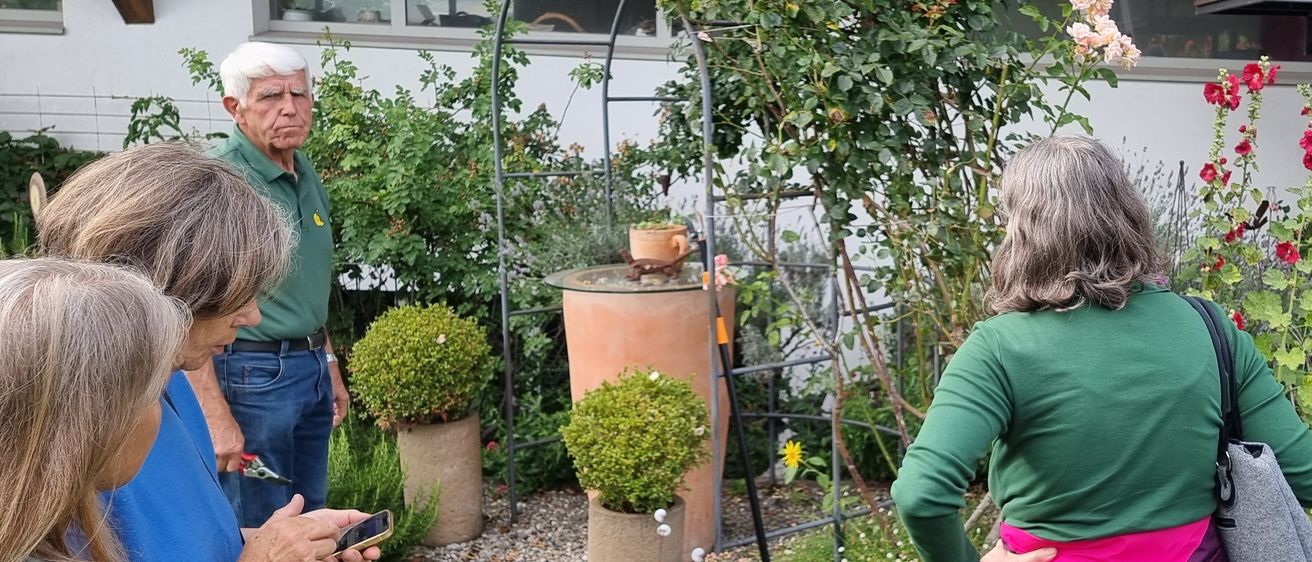 Eine Gruppe von Menschen ist draußen in einem Garten. Eine Person hält ein Telefon und schaut nach unten. Ein älterer Mann steht neben einer Frau, die die Hände auf die Hüften gestemmt hat.