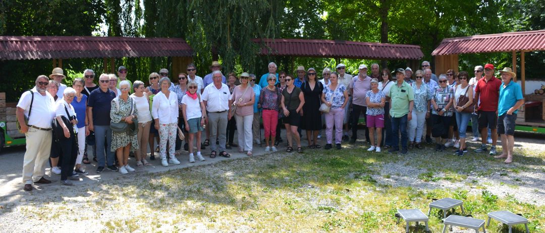 Eine Gruppe von Menschen in Sonnenbrillen und Hüten steht vor einem Gebäude mit einem roten Dach. Dahinter sind Bäume und Gras.