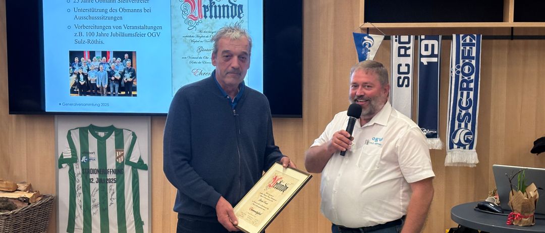 Zwei Männer stehen vor einer Holzwand, einer hält eine Auszeichnung und ein Mikrofon. Der Mann im weißen Hemd spricht in das Mikrofon. Hinter ihnen ist eine Projektionsleinwand mit einer Folie. Ein grün-weiß gerahmtes Trikot hängt an der Wand.