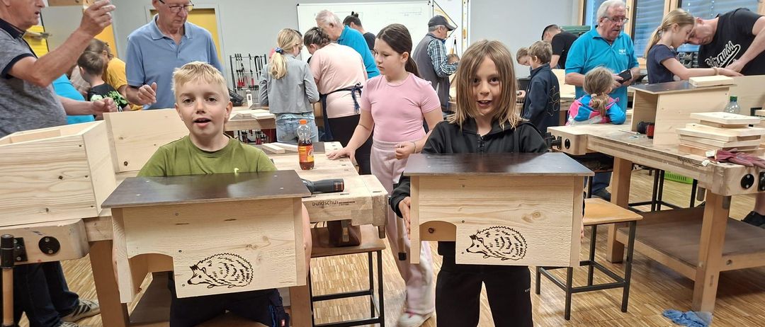 Kinder in einer Werkstatt, die Holzkisten mit Igel-Designs halten, während andere an Schreibtischen arbeiten. Ein älterer Mann steht hinter ihnen.