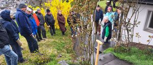 Eine Gruppe von Menschen versammelt sich in einem Garten. Ein Mann kniet mit einer Schaufel. Andere stehen und lauschen einem Redner. Pflanzen mit gelben Blumen und Grün umgeben sie.