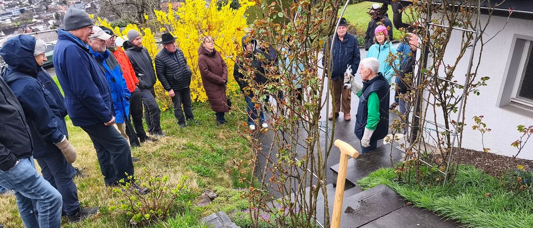 Eine Gruppe von Menschen versammelt sich in einem Garten. Ein Mann kniet mit einer Schaufel. Andere stehen und lauschen einem Redner. Pflanzen mit gelben Blumen und Grün umgeben sie.