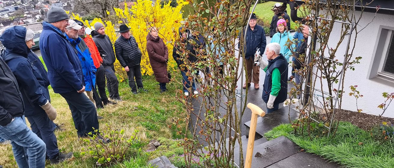 Eine Gruppe von Menschen versammelt sich in einem Garten. Ein Mann kniet mit einer Schaufel. Andere stehen und lauschen einem Redner. Pflanzen mit gelben Blumen und Grün umgeben sie.