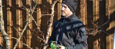 Ein Mann in einer schwarzen Jacke und Mütze, mit Gartenschere in der Hand, steht neben einem Holzzaun mit einem Baumzweig.