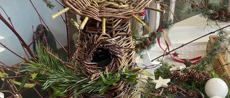 Ein handgefertigtes Vogelhaus aus Zweigen und Grünpflanzen mit einem kleinen Loch steht auf einem Betonsockel neben einem Weihnachtskranz und Tannenzapfen.