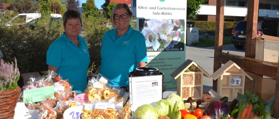 Zwei Frauen stehen vor einem Stand mit verschiedenen Obst und Gemüse. Sie tragen blaue Hemden mit dem Logo OGV und sind von Pflanzen und einem Schild umgeben.