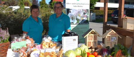 Two women stand in front of a booth with various fruits and vegetables. They wear blue shirts with the logo OGV and are surrounded by plants and a sign.