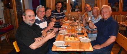 Eine Gruppe von Männern sitzt drinnen um einen Tisch, lächelt und hält Gläser mit Bier in die Höhe. Sie sind umgeben von Tellern, Weingläsern und Essen. Der Raum hat große Fenster mit Holzrahmen.