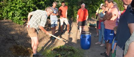 Eine Gruppe von Männern auf einem Feldweg. Ein Mann benutzt eine Heugabel zum Graben, während andere zusehen. Ein blauer Eimer und ein grünes Objekt sind in der Nähe.