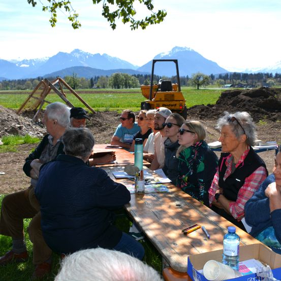 Eine Gruppe von Menschen sitzt auf einem Holztisch in einem Grasfeld und diskutiert bei einem Blick auf die Berge im Hintergrund.