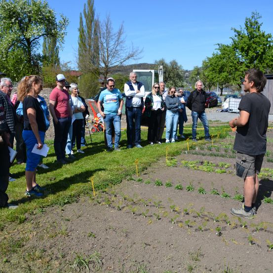 Eine Gruppe von Menschen steht in einem Feld und hört einem Mann zu, der etwas erklärt. Der Mann hält ein Stück Papier und zeigt auf etwas im Boden.