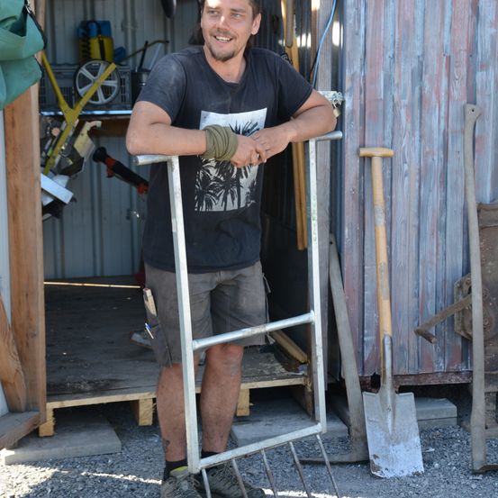 Ein Mann steht in einer Scheune und lehnt sich mit verschränkten Armen an eine Metallrechen, lächelt. Die Scheune ist mit verschiedenen Werkzeugen übersät, darunter eine Schaufel und ein Rechen.
