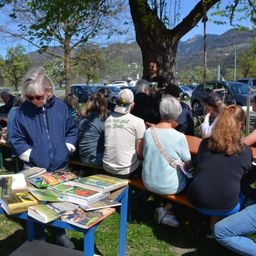 Eine Gruppe von Menschen, wahrscheinlich ein Buchclub, sitzt an einem Tisch voller Bücher an einem sonnigen Tag, mit einem Mann, der hinter ihnen steht, und einem Baum im Hintergrund.