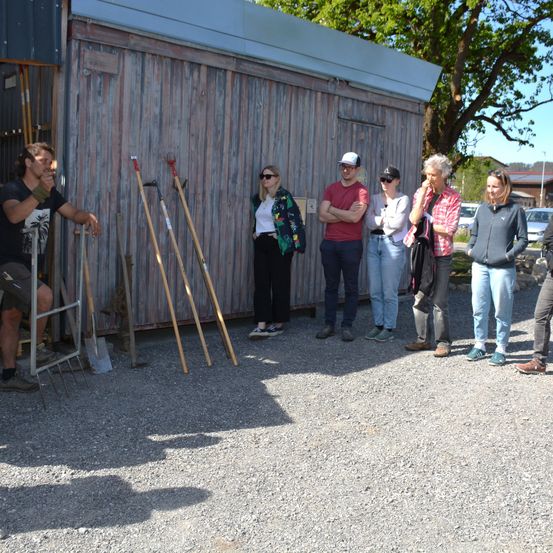 Eine Gruppe von Menschen steht vor einem Gebäude, einige mit verschränkten Armen. Ein Mann sitzt auf einer Leiter und hält eine Harke. Ein Baum steht hinter dem Gebäude, und Autos sind in der Nähe geparkt.
