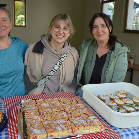 Drei Frauen stehen hinter einem Tisch mit einer Tablett voller Desserts. Die Frau auf der linken Seite trägt ein blaues T-Shirt, die Frau in der Mitte trägt eine graue Kapuzenjacke mit einem weißen Riemen und die Frau auf der rechten Seite trägt eine grüne Jacke.