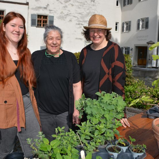 Drei Frauen vor einem Gebäude mit Pflanzen und einem Holztisch. Die Frau links hat rote Haare und eine braune Jacke. Die Frau in der Mitte hat graues Haar, ein schwarzes Sweatshirt und eine Brille. Die Frau rechts trägt einen Hut und ein schwarzes Sweatshirt.