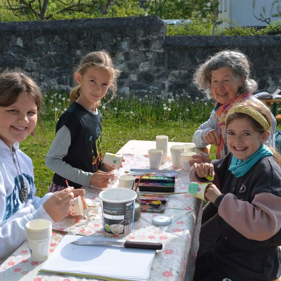 Vier Mädchen sitzen an einem Tisch mit Tassen und Farbe. Sie bemalen eine Tasse mit einem Blumendesign.