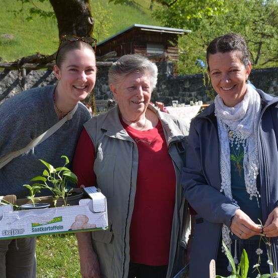 Drei Frauen stehen nebeneinander auf einem Grasfeld. Die Frau auf der linken Seite hält eine Schachtel mit Pflanzen. Die Frau in der Mitte trägt eine Halskette und ein rotes Shirt. Die Frau auf der rechten Seite trägt eine graue Jacke und eine Schürze.
