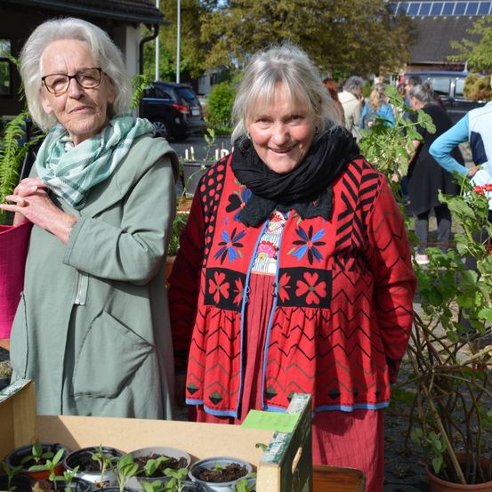 Zwei ältere Frauen stehen nebeneinander in einem Garten. Eine trägt einen grünen Mantel, die andere ein rotes Kleid mit schwarzem Schal.