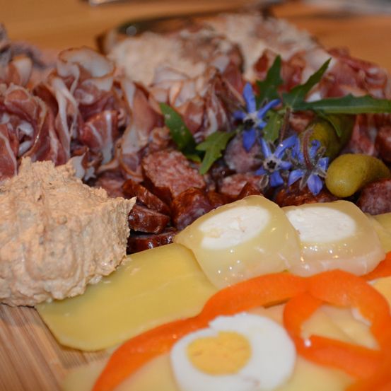 Ein Holzschneidebrett ist mit verschiedenen Wurstsorten, Käsescheiben, einer Scheibe gekochtes Ei und Orangen-Paprika-Scheiben belegt.