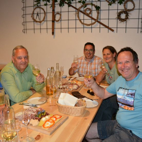 Eine Gruppe Erwachsener sitzt an einem Tisch mit Weinläsern, Essen und Flaschen und lächelt.