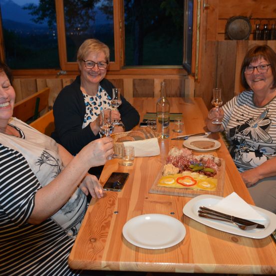 Drei Frauen sitzen an einem Holztisch, lächeln und halten Weingläser. Vor ihnen liegt ein Käsetablett, Brot und verschiedene Getränke.