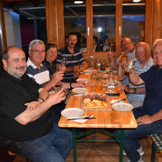 Eine Gruppe von Erwachsenen sitzt an einem Tisch, hält Gläser mit Wein und Bier in den Händen und lächelt für ein Foto.