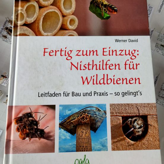 Ein Buch mit dem Titel 'Fertig zum Einzug: Nisthilfen fur Wildbienen'. Es zeigt Bilder von Bienen, Bienennestern und einer Biene auf einem Blatt. Der Text deutet darauf hin, dass es sich um einen Leitfaden für den Bau und die Praxis mit Wildbienen handelt.