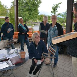 Eine Gruppe von Menschen steht um einen Tisch mit verschiedenen Gegenständen. Ein Mann hält eine Holzkiste, während eine Frau ein Blatt Papier liest.