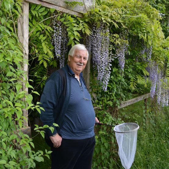 Ein älterer Mann steht in einem Garten, lächelt und hält ein Netz, umgeben von Pflanzen mit lila Blüten.