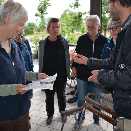 Eine Gruppe älterer Personen steht in einem Garten. Eine Frau in einer blauen Jacke hält einen Prospekt und schaut einen Mann in einer schwarzen Jacke an, der ein kleines Glas hält. Hinter ihnen beobachten andere Personen die Interaktion.
