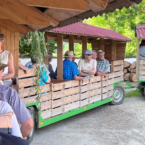 Eine Gruppe von Menschen fährt auf einem hölzernen Wagen, trägt Hüte und Brillen. Der Wagen hat ein rotes Dach und steht in einem Hof mit einem Holzzaun.