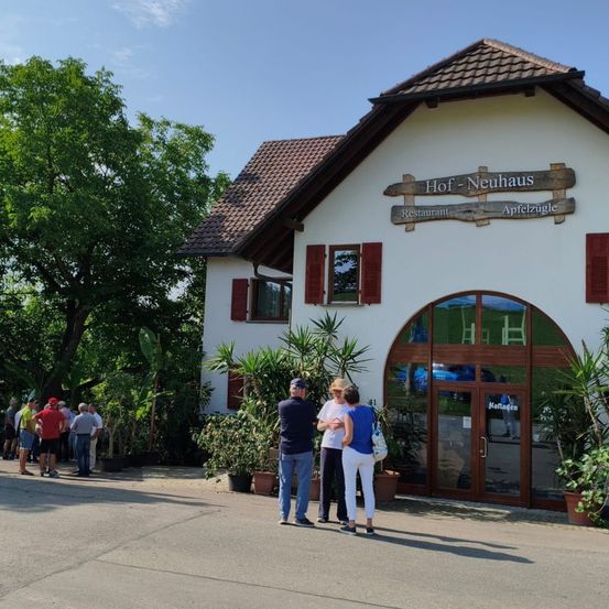 Menschen versammeln sich vor dem Restaurant und Apfelorchard Hof-Neuhaus. Das Gebäude hat ein Schild mit der Aufschrift 'Hof-Neuhaus', darunter 'Restaurant' und 'Apfelzugle'. Im Vordergrund stehen Topfpflanzen und ein Baum.