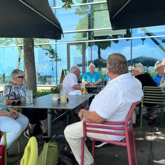 Eine Gruppe älterer Personen sitzt an einem Tisch vor einem Restaurant. Sie führen ein Gespräch, und auf dem Tisch stehen Gläser mit Getränken. Die Außenumgebung ist mit grünen Pflanzen und Stühlen geschmückt.